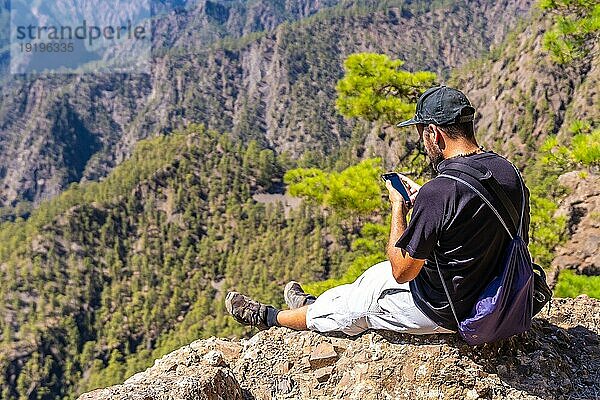 Ein junger Mann ruht sich nach einer Wanderung auf dem Gipfel des La Cumbrecita aus  sitzt auf dem natürlichen Aussichtspunkt und macht ein Foto mit dem Handy  Insel La Palma  Kanarische Inseln  Spanien  Europa