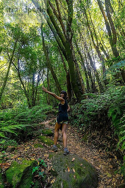 Eine glückliche junge Frau  die im Naturpark Cubo de la Galga an der Nordostküste der Insel La Palma (Kanarische Inseln) einen Wanderweg neben Farnen beschreitet. Spanien