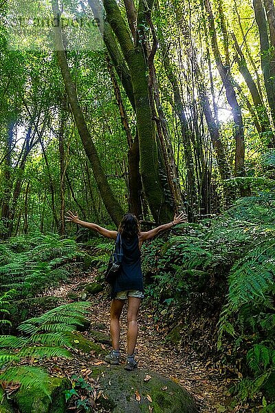 Eine glückliche junge Frau  die im Naturpark Cubo de la Galga an der Nordostküste der Insel La Palma (Kanarische Inseln) einen Wanderweg neben Farnen beschreitet. Spanien