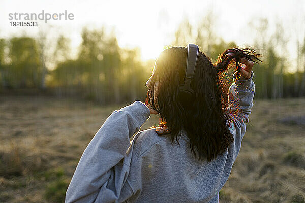 Rückansicht einer Frau  die bei Sonnenuntergang auf einer Wiese Musik hört