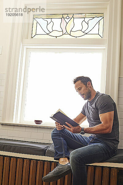 Mann liest Buch und sitzt zu Hause am Fenster