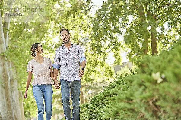 Lächelndes Paar hält sich an den Händen und geht auf einem von Bäumen gesäumten Gehweg