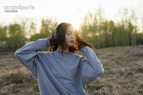 Porträt einer Frau  die bei Sonnenuntergang auf einer Wiese mit geschlossenen Augen Musik hört