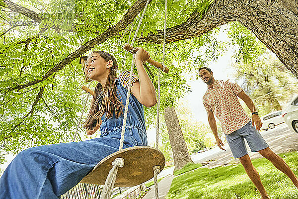 Lächelndes Mädchen (12-13) auf Schaukel im Park  Vater im Hintergrund