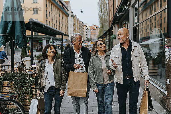 Lächelnde ältere Freunde halten Einkaufstüten in der Hand und reden  während sie auf dem Bürgersteig gehen