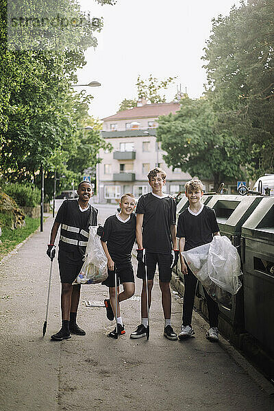 Ganzkörperporträt lächelnder Jungen  die mit Müllsäcken neben Recyclingbehältern stehen