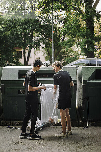 Ganzkörperansicht von Jungen im Teenageralter  die einen Müllsack mit Plastik neben der Recyclingtonne halten