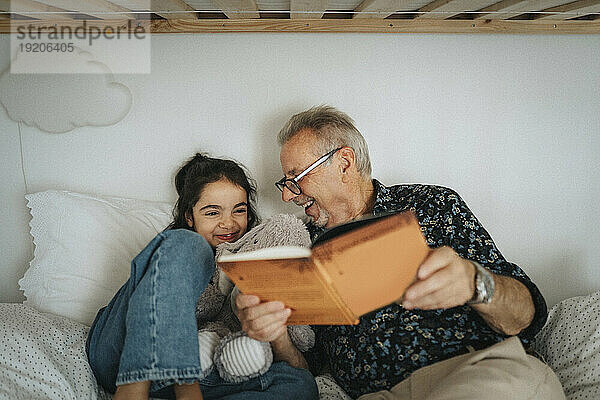 Glücklicher älterer Mann  der zu Hause im Bett mit seiner Enkelin ein Märchenbuch liest