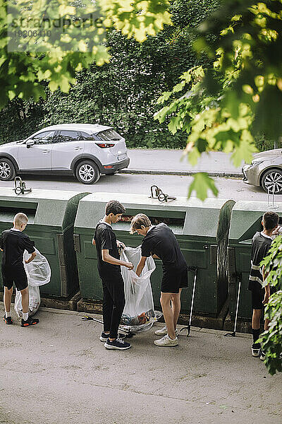 Freiwillige in voller Länge halten einen Müllsack mit Plastik neben der Recyclingtonne