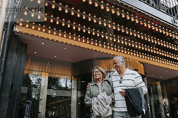 Happy senior man with arm around female elderly friend while standing outside movie theater