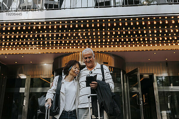 Lächelnder älterer Mann  der sein Smartphone mit einer älteren Freundin vor dem Kino teilt