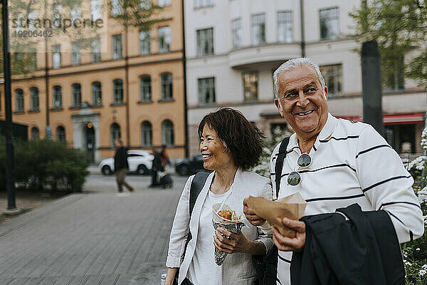 Lächelnde männliche und weibliche ältere Freunde schauen weg  während sie auf der Straße Snacks in der Hand halten