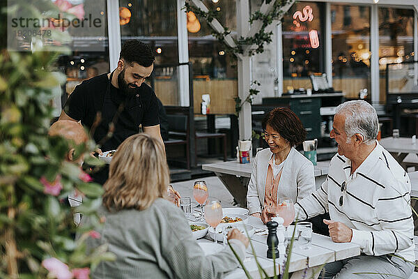 Glückliche männliche und weibliche ältere Freunde schauen dem Kellner zu  der im Restaurant Essen serviert