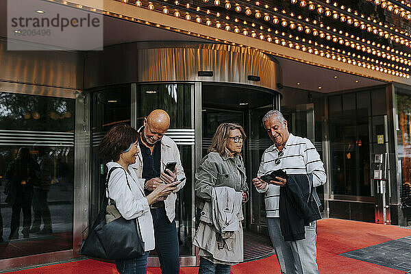 Männliche und weibliche ältere Freunde teilen Smartphones  während sie vor dem Kino stehen