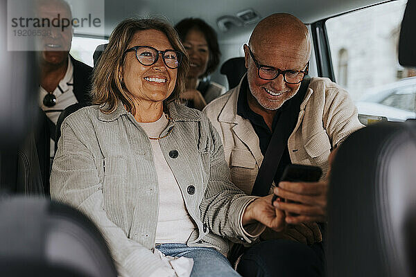 Glückliche männliche und weibliche ältere Freunde  die ihr Smartphone teilen  während sie im Auto sitzen
