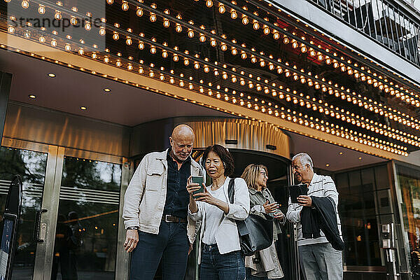 Männliche und weibliche ältere Freunde diskutieren miteinander  während sie Smartphones vor dem Kino benutzen