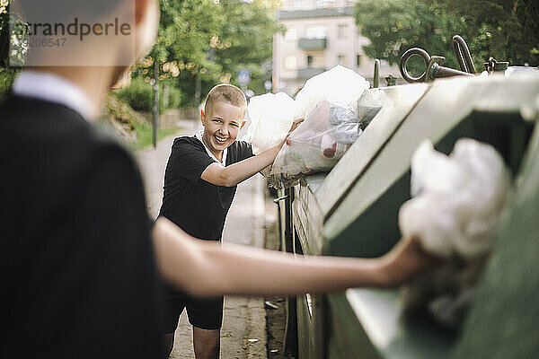 Lächelnder Junge schaut einen Freund an  während er Müllsäcke in Recyclingbehälter wirft