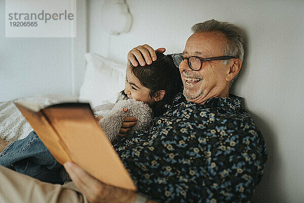 Fröhlicher älterer Mann liest mit Enkelin im heimischen Bett ein Bilderbuch