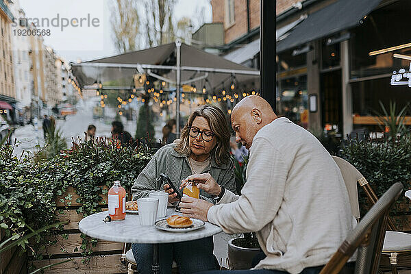Männliche und weibliche ältere Freunde diskutieren über das Smartphone  während sie im Straßencafé etwas essen