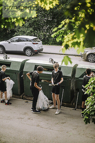 Ganzkörperansicht von Jungen  die einen Müllsack mit Plastik neben der Recyclingtonne halten