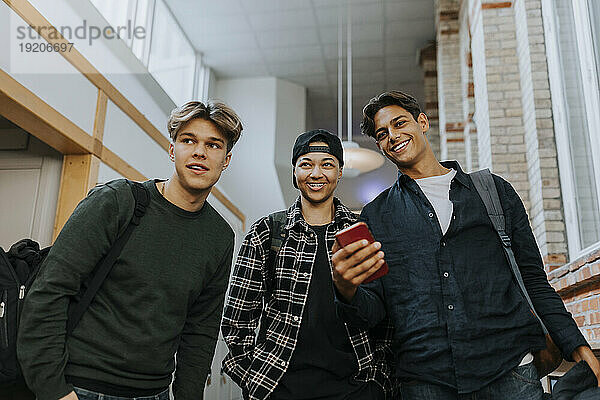 Low-Angle-Ansicht lächelnder männlicher Freunde mit Mobiltelefon beim Spaziergang durch den Schulkorridor