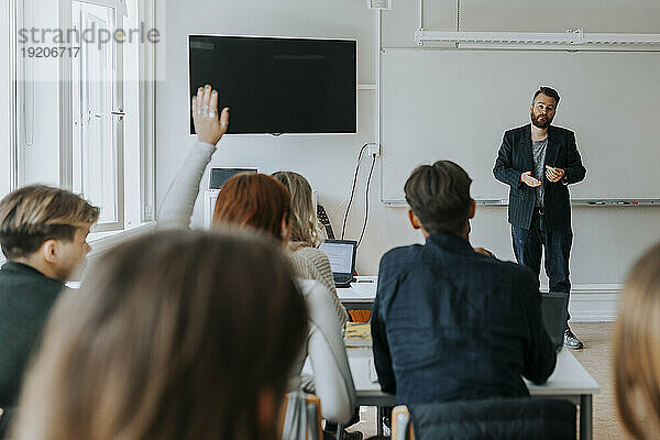 Ein männlicher Professor unterrichtet  während eine Studentin mit erhobener Hand im Klassenzimmer sitzt