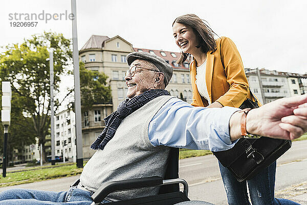 Lachende junge Frau schiebt glücklichen älteren Mann im Rollstuhl