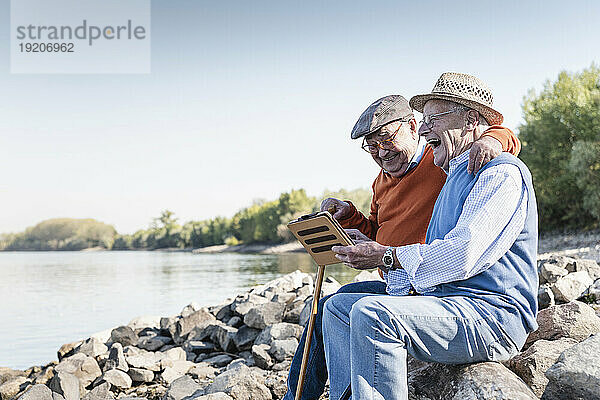 Zwei alte Freunde sitzen am Flussufer und nutzen ein digitales Tablet