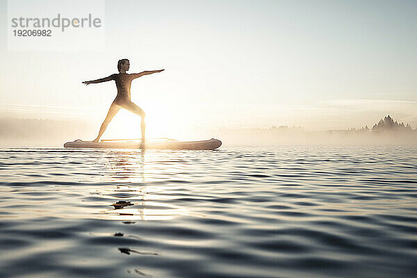 Frau praktiziert Paddle-Board-Yoga am Kirchsee am Morgen  Bad Tölz  Bayern  Deutschland