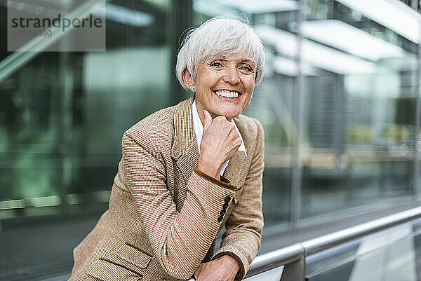 Porträt einer lächelnden älteren Geschäftsfrau  die sich in der Stadt auf ein Geländer stützt