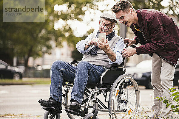 Lachender älterer Mann im Rollstuhl und sein erwachsener Enkel schauen gemeinsam aufs Smartphone und haben Spaß