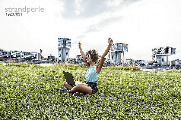 Deutschland  Köln  jubelnde Frau sitzt mit Laptop auf einer Wiese