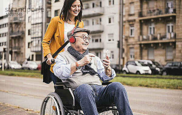 Lächelnde junge Frau schiebt einen glücklichen älteren Mann mit Kopfhörern und Smartphone im Rollstuhl