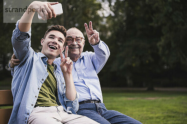 Älterer Mann und Enkel sitzen zusammen auf einer Parkbank und machen ein Selfie mit dem Smartphone