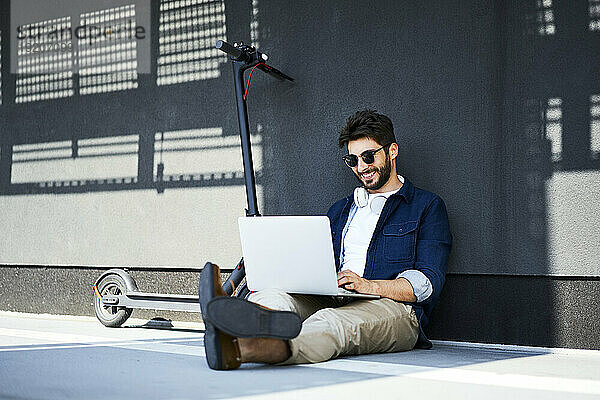 Lächelnder junger Mann  der neben seinem Elektroroller auf dem Boden sitzt und einen Laptop benutzt