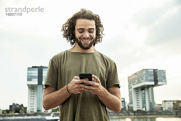 Deutschland  Köln  lächelnder junger Mann  der auf sein Handy schaut
