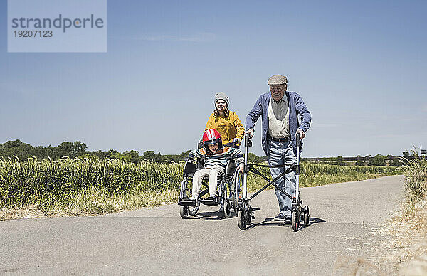 Mädchen mit Jungen sitzen im Rollstuhl und älterer Mann hält Gehhilfe auf der Straße