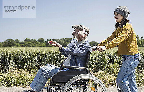 Lächelndes Mädchen mit Großvater  der an einem sonnigen Tag im Rollstuhl sitzt und Musik hört