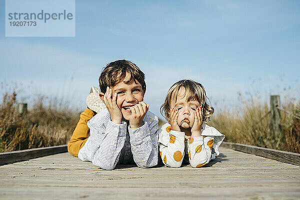 Porträt eines Jungen und seiner kleinen Schwester  die Seite an Seite auf der Promenade liegen und lustige Gesichter ziehen