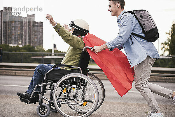 Junger Mann schiebt älteren Mann im Rollstuhl  der als Superheld verkleidet ist