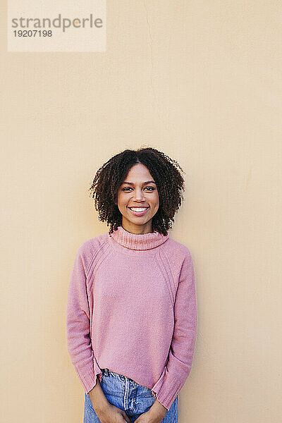 Porträt einer lächelnden jungen Frau in rosa Pullover  die an der Wand lehnt