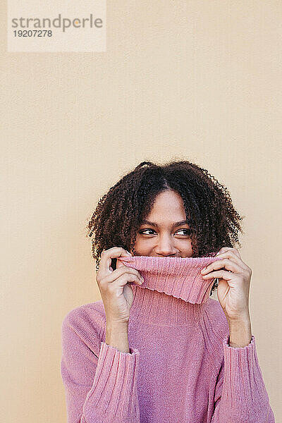 Portrait of smiling young woman wearing pink turtleneck pullover leaning against wall