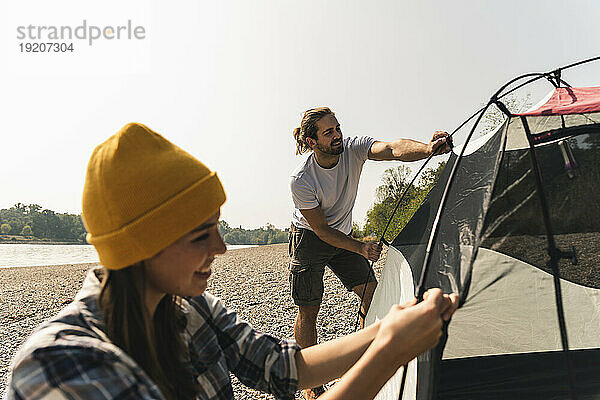 Lächelndes junges Paar baut am Flussufer ein Zelt auf