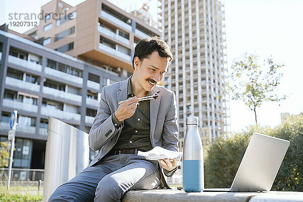 Geschäftsmann isst zu Mittag und telefoniert per Laptop im Büropark