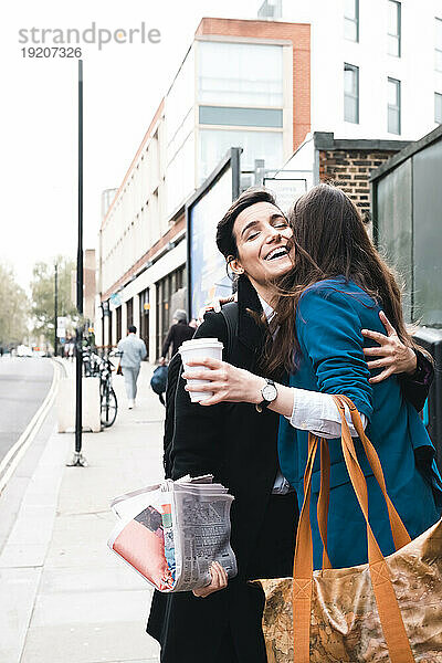 Glückliche Geschäftsfrau umarmt Freundin mit Kaffeetasse am Bürgersteig