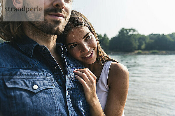 Lächelnde junge Frau lehnt am Flussufer an der Schulter eines Mannes