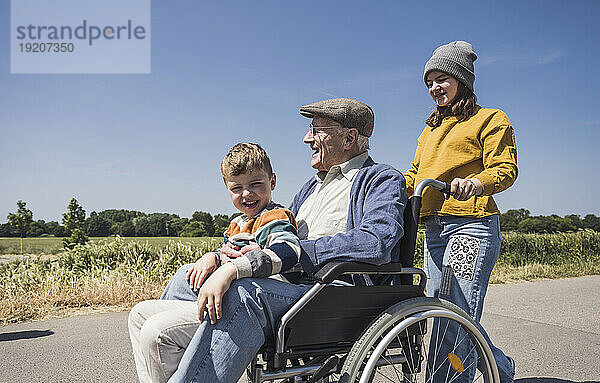 Lächelndes Mädchen mit Großvater und Enkel sitzen an einem sonnigen Tag im Rollstuhl