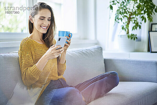Lächelnde junge Frau sitzt mit Smartphone und einer Tasse Kaffee auf dem Sofa