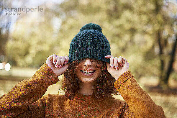 Cheerful redhead woman covering eyes with knit hat at autumn park