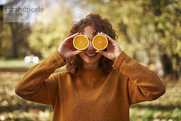 Smiling redhead woman holding orange slices in front of face at autumn park
